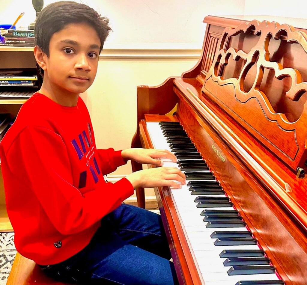Boy in an orange sweatshirt smiling at the camera while sitting at the piano