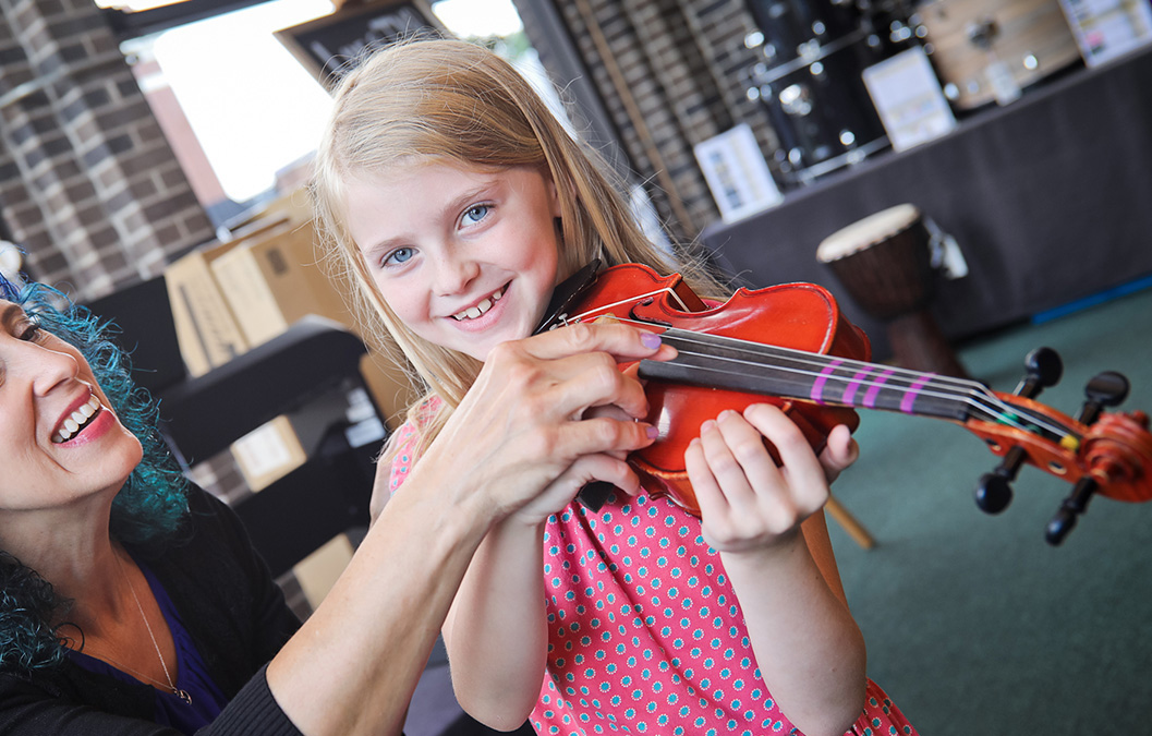 Young girl holding a violin and smiling at camera, teacher is helping her hold her hands correctly