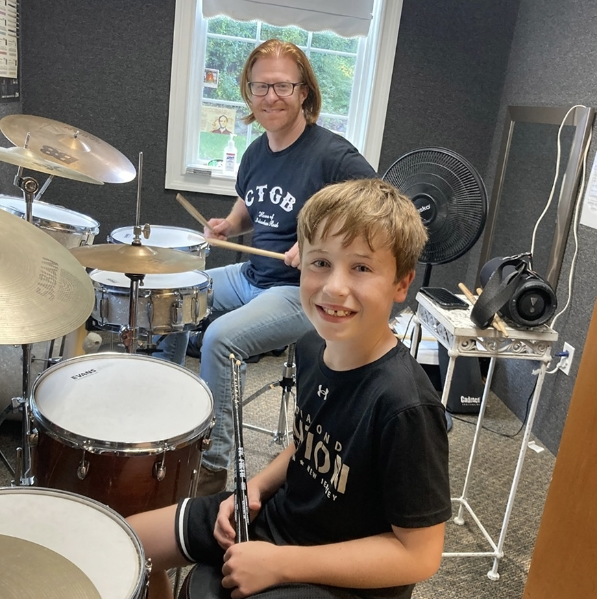 Male drum teacher with young male student both are smiling at the camera