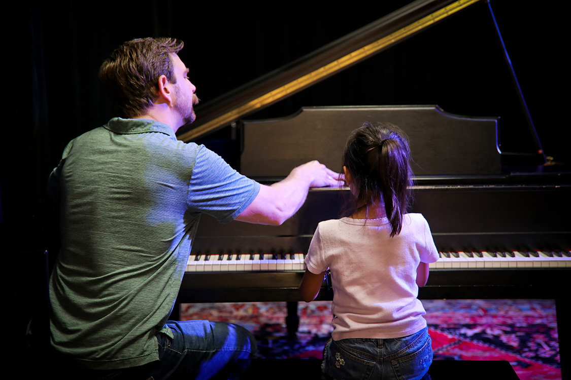 Male music teacher sitting at a piano instructing a young girl seated beside him