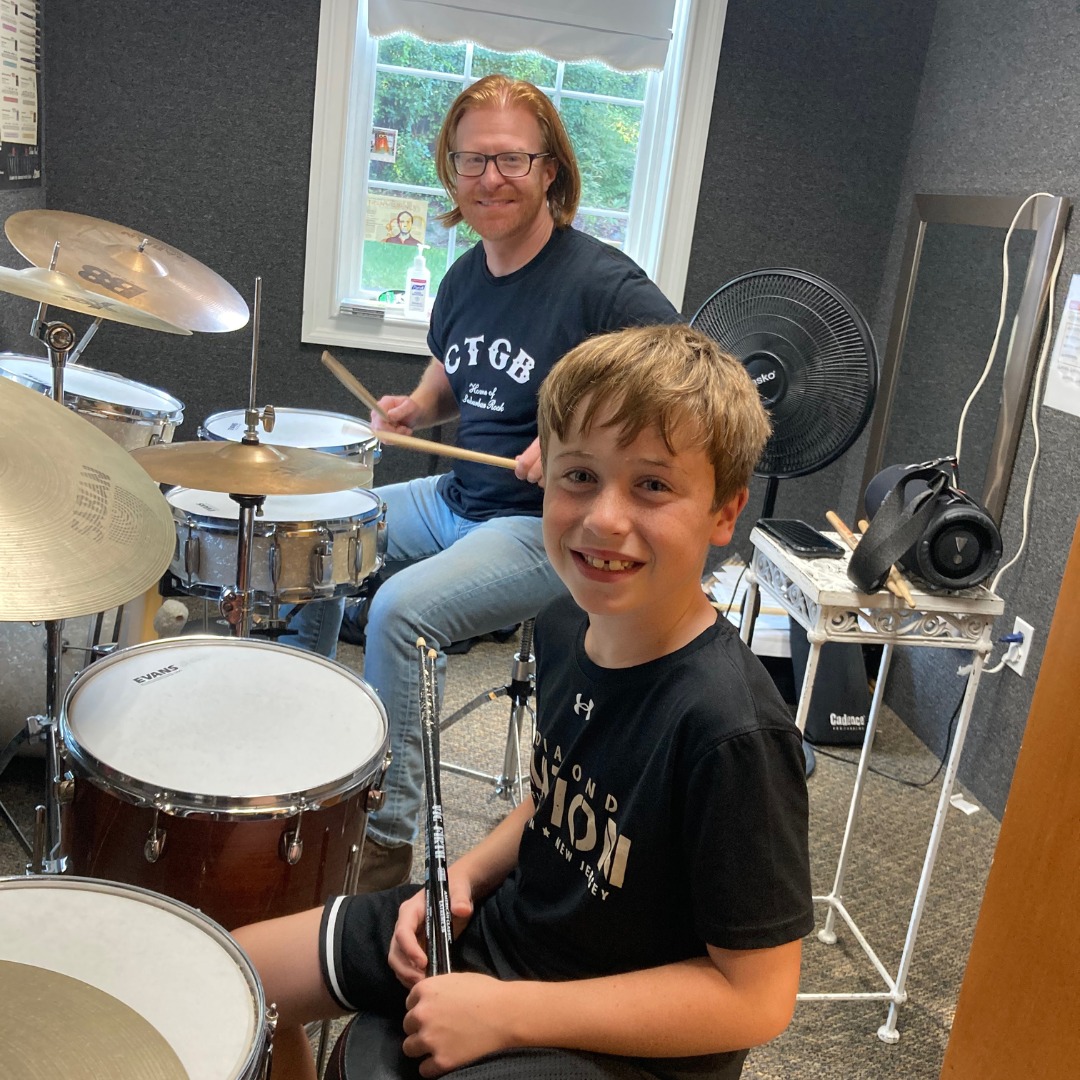 Male drum teacher with young male student both are smiling at the camera