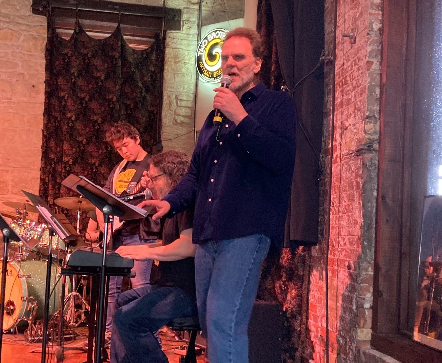male instructor on stage singing next to a piano playing teacher and student playing guitar