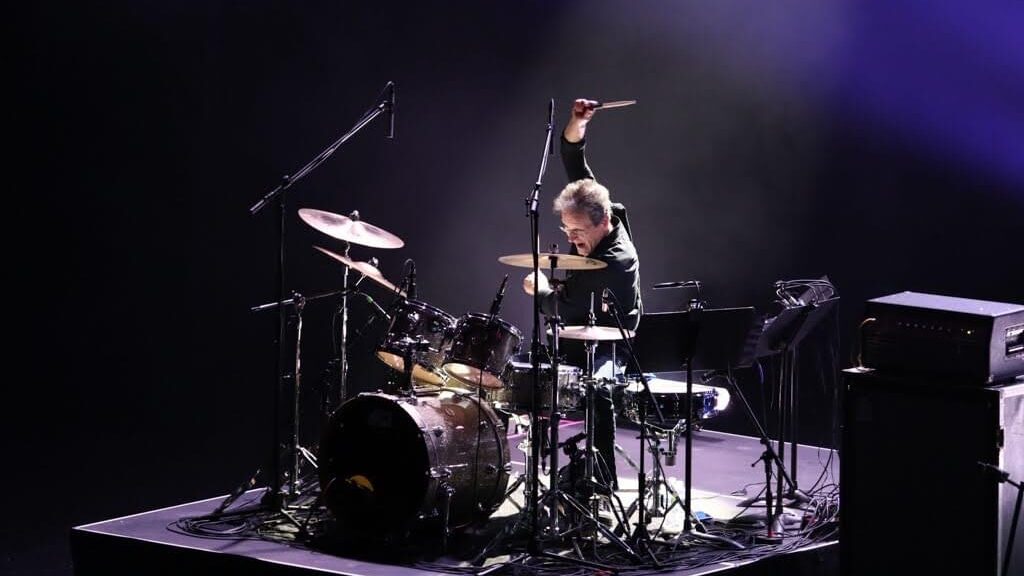 drum instructor playing on stage with his stick raised up against a dark background