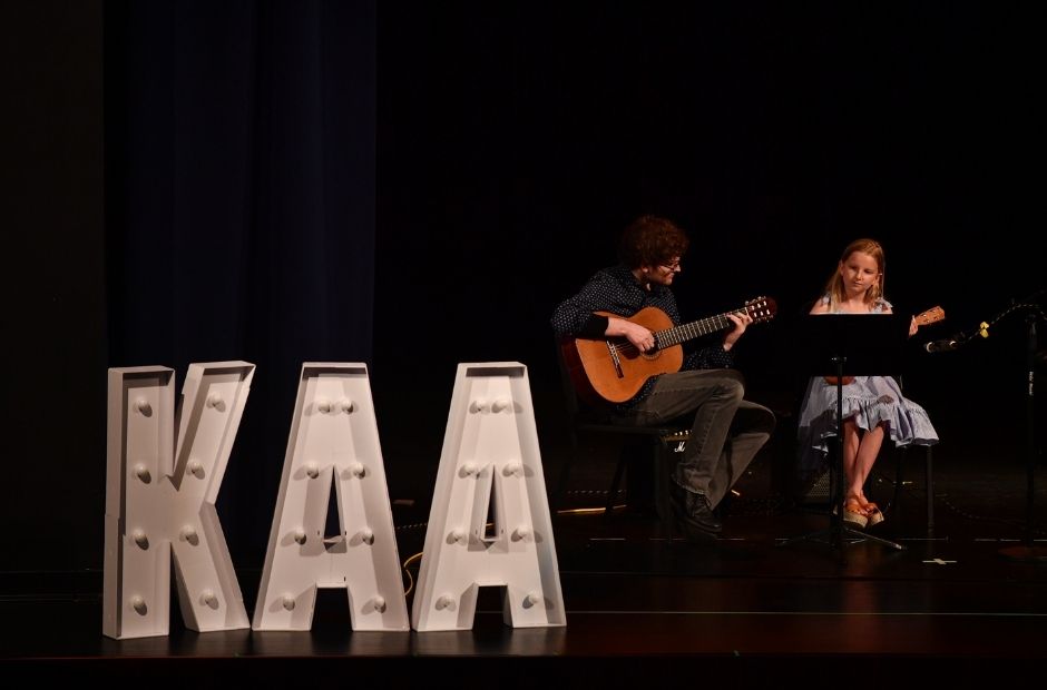 students playing guitar at a recitall with a kentucky Arts Academy marquee sitting at the front of the stage