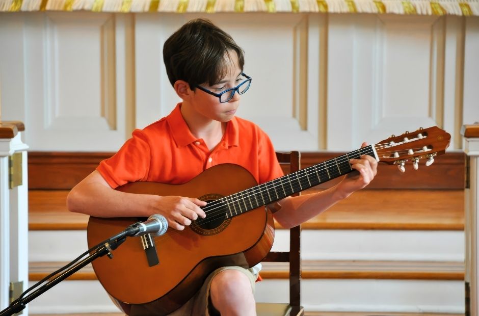 guitarist performing in a recital in an orange shirt and green glasses holding an acoustic guitar