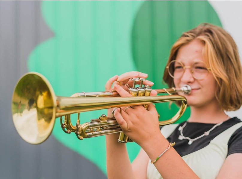 Young girl with short blonde hair and glasses playing a trumpet.