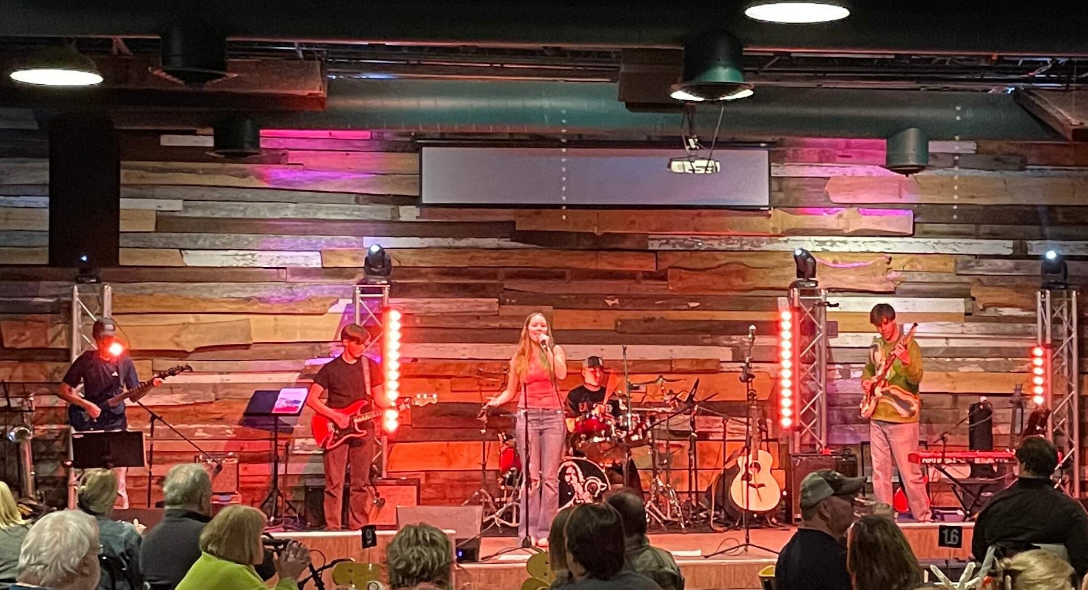 a photo of one of our student bands playing on stage. There's a wooden slat background on the wall and red lights. There's a drummer, singer, 2 electric guitarists and a bassist performing in front of a large crowd