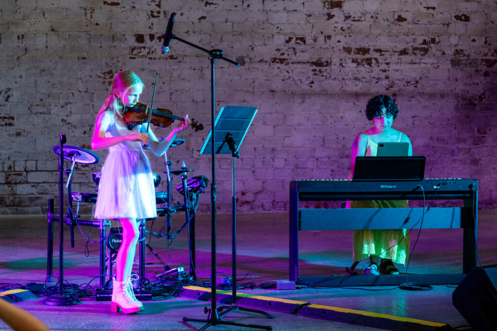 Girl on stage playing violin with teacher accompanying on piano