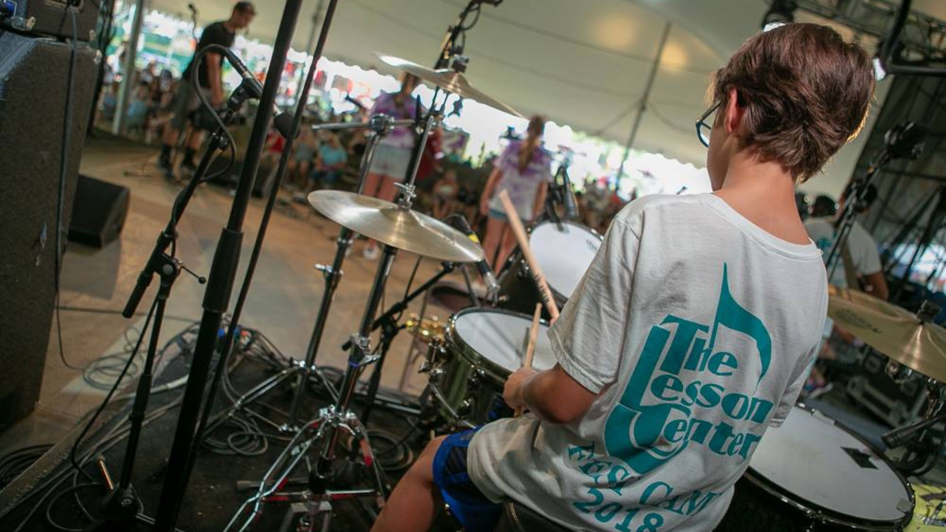Photograph of a boy on a stage playing drumset from behind wearing a The Lesson Center shirt