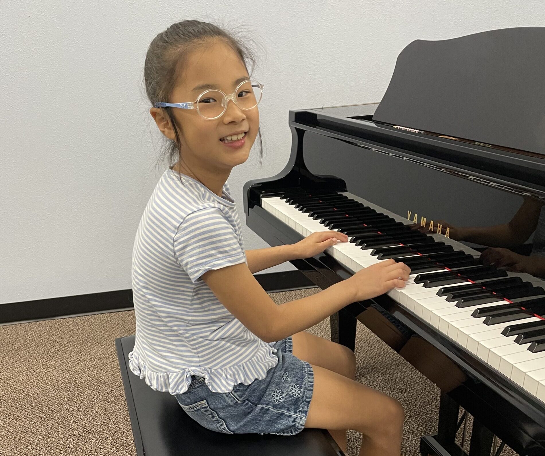 young girl sitting at a piano, smiling at the camera wearing glasses