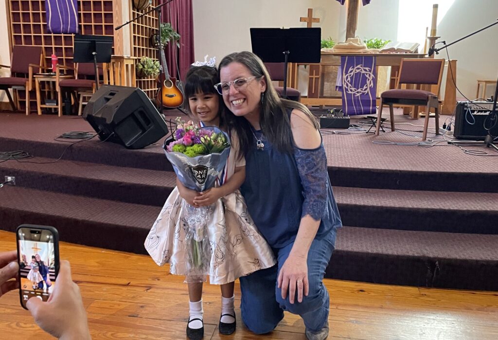 teacher and young student posing in front of the stage after a recital. little girl student is holding a bouquet of flowers