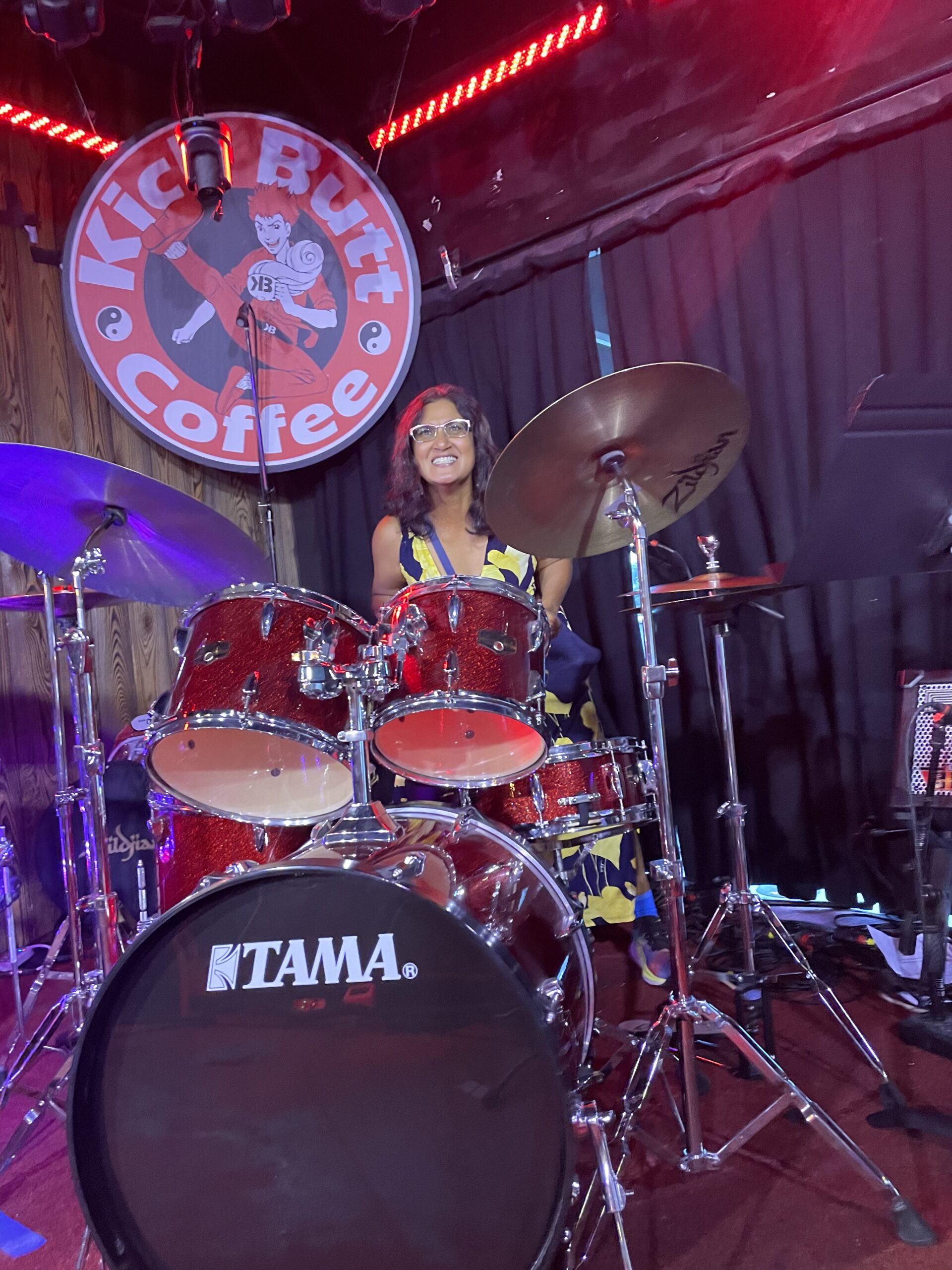 a woman wearing glasses, sitting behind a red TAMA drum set on stage.